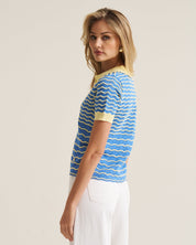 A woman modeling a short-sleeve knit top with a blue and white wavy stripe pattern and yellow collar/cuff trim, paired with white pants, against a plain light beige studio background.