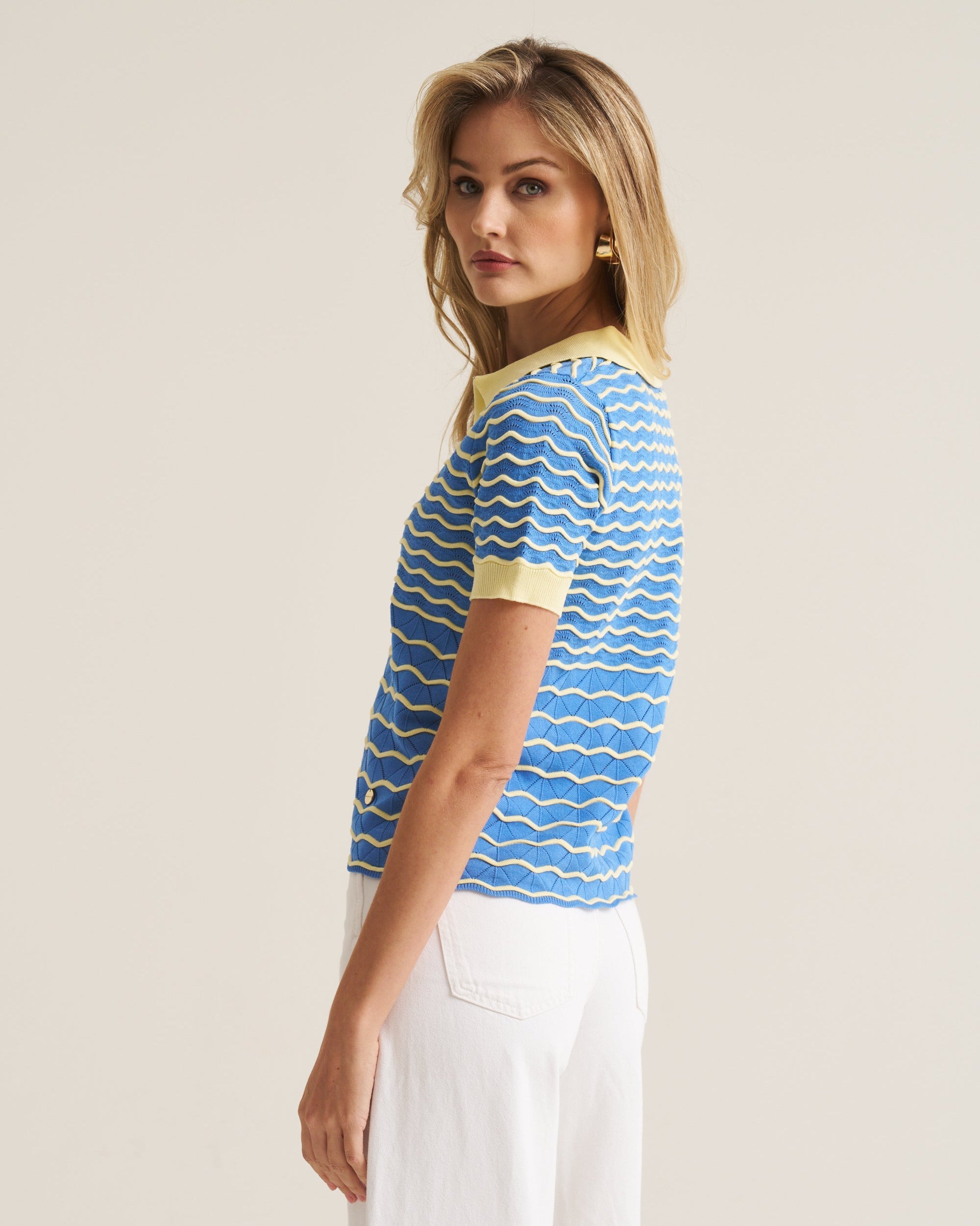 A woman modeling a short-sleeve knit top with a blue and white wavy stripe pattern and yellow collar/cuff trim, paired with white pants, against a plain light beige studio background.