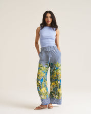 A woman modeling wide-leg pants with a blue and white striped upper section and a vibrant blue, green, and yellow tropical print lower section, paired with a light blue sleeveless top and brown sandals, against a plain beige studio background.