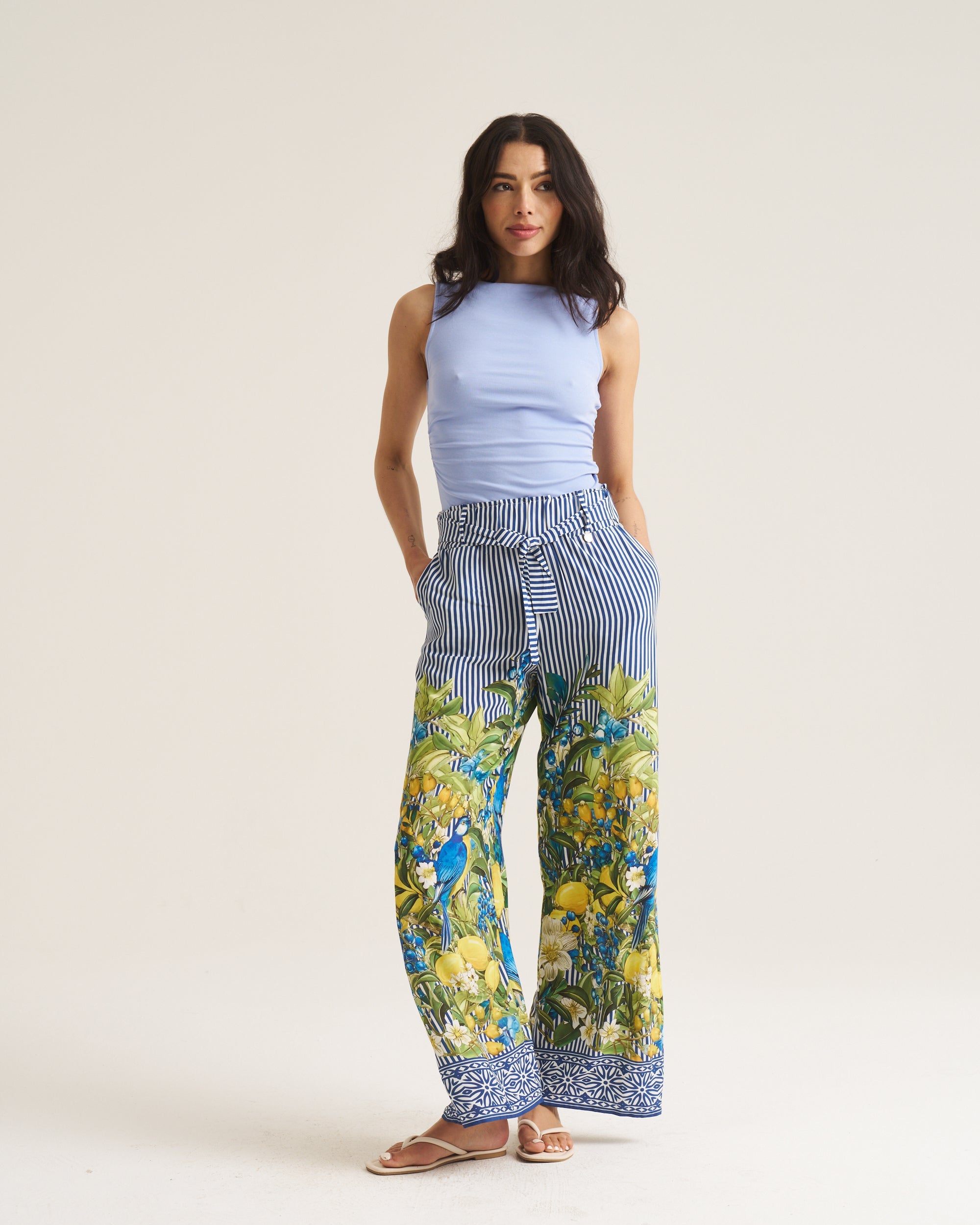A woman modeling wide-leg pants with a blue and white striped upper section and a vibrant blue, green, and yellow tropical print lower section, paired with a light blue sleeveless top and brown sandals, against a plain beige studio background.