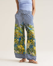 A person wearing wide-leg pants with a blue and white vertical stripe pattern on the upper portion and a colorful botanical print with blue birds and yellow fruit on the lower legs, paired with a light blue top and tan sandals, against a plain light beige studio background.