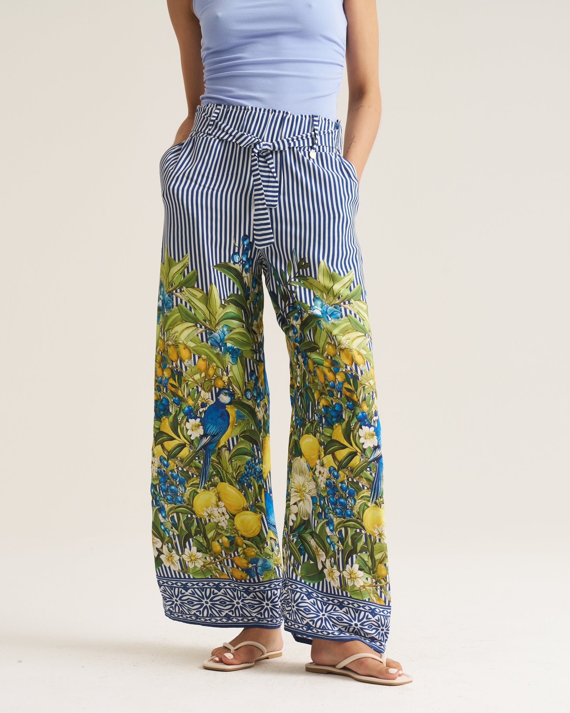 A person wearing wide-leg pants with a blue and white vertical stripe pattern on the upper portion and a colorful botanical print with blue birds and yellow fruit on the lower legs, paired with a light blue top and tan sandals, against a plain light beige studio background.