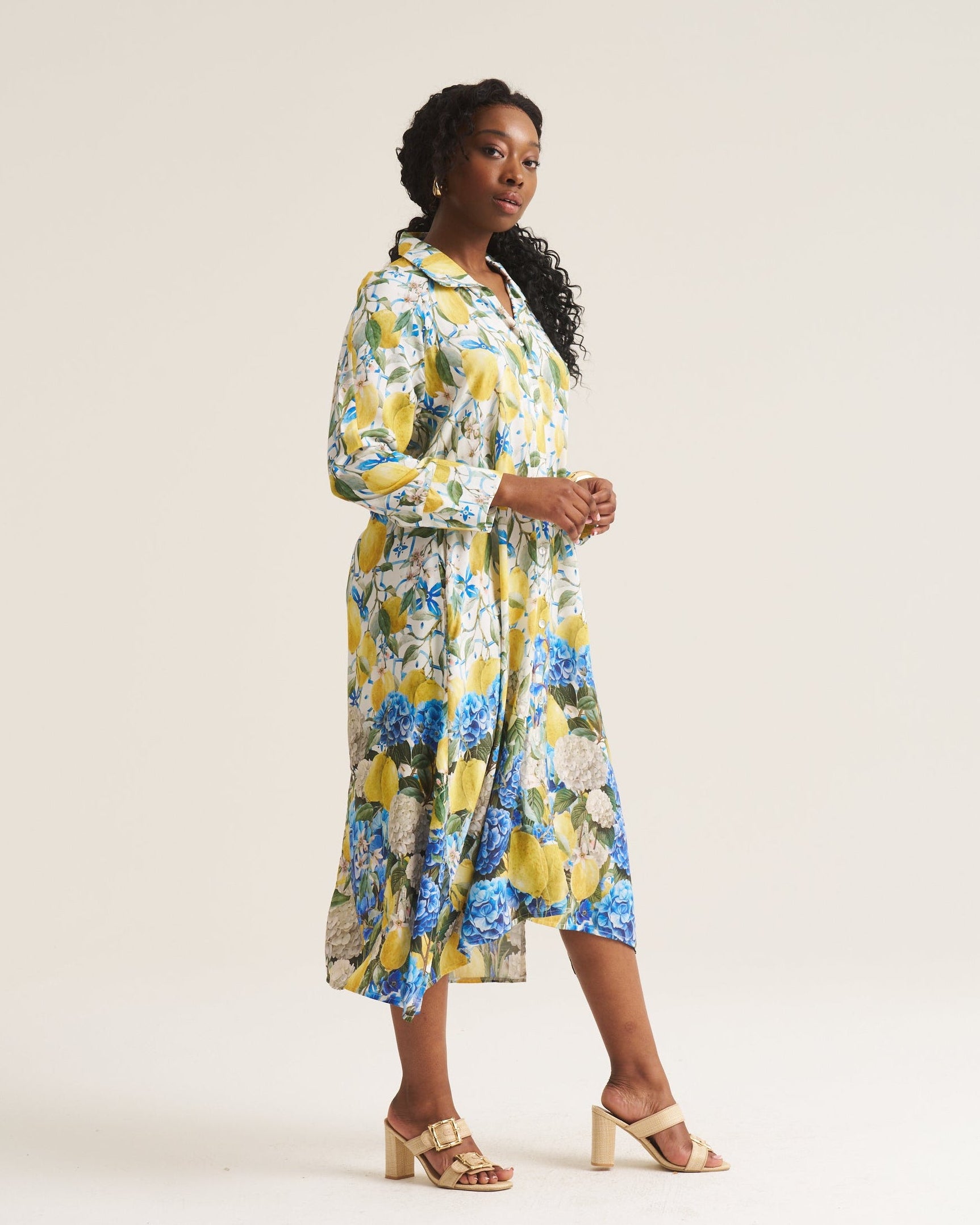 A woman modeling a long-sleeve midi shirt dress with a vibrant yellow, blue, and white floral print, paired with beige block-heel sandals, against a plain off-white studio background.