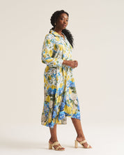 A woman modeling a long-sleeve midi shirt dress with a vibrant yellow, blue, and white floral print, paired with beige block-heel sandals, against a plain off-white studio background.