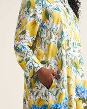 A close-up of a person wearing a long-sleeve dress with a vibrant lemon and blue hydrangea floral print on a cream background, featuring a side pocket.