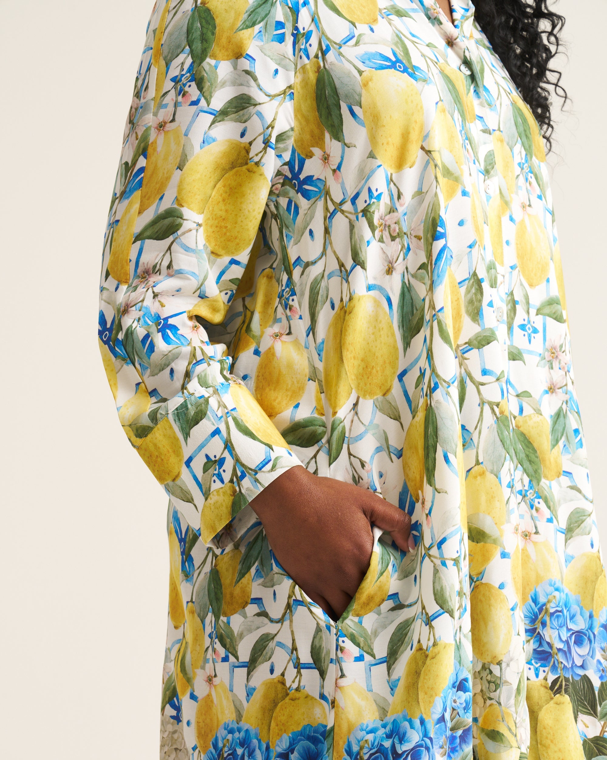 A close-up of a person wearing a long-sleeve dress with a vibrant lemon and blue hydrangea floral print on a cream background, featuring a side pocket.