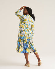 A woman modeling a long-sleeved midi dress with a vibrant yellow, blue, and white floral print, photographed from behind against a solid light beige studio background.