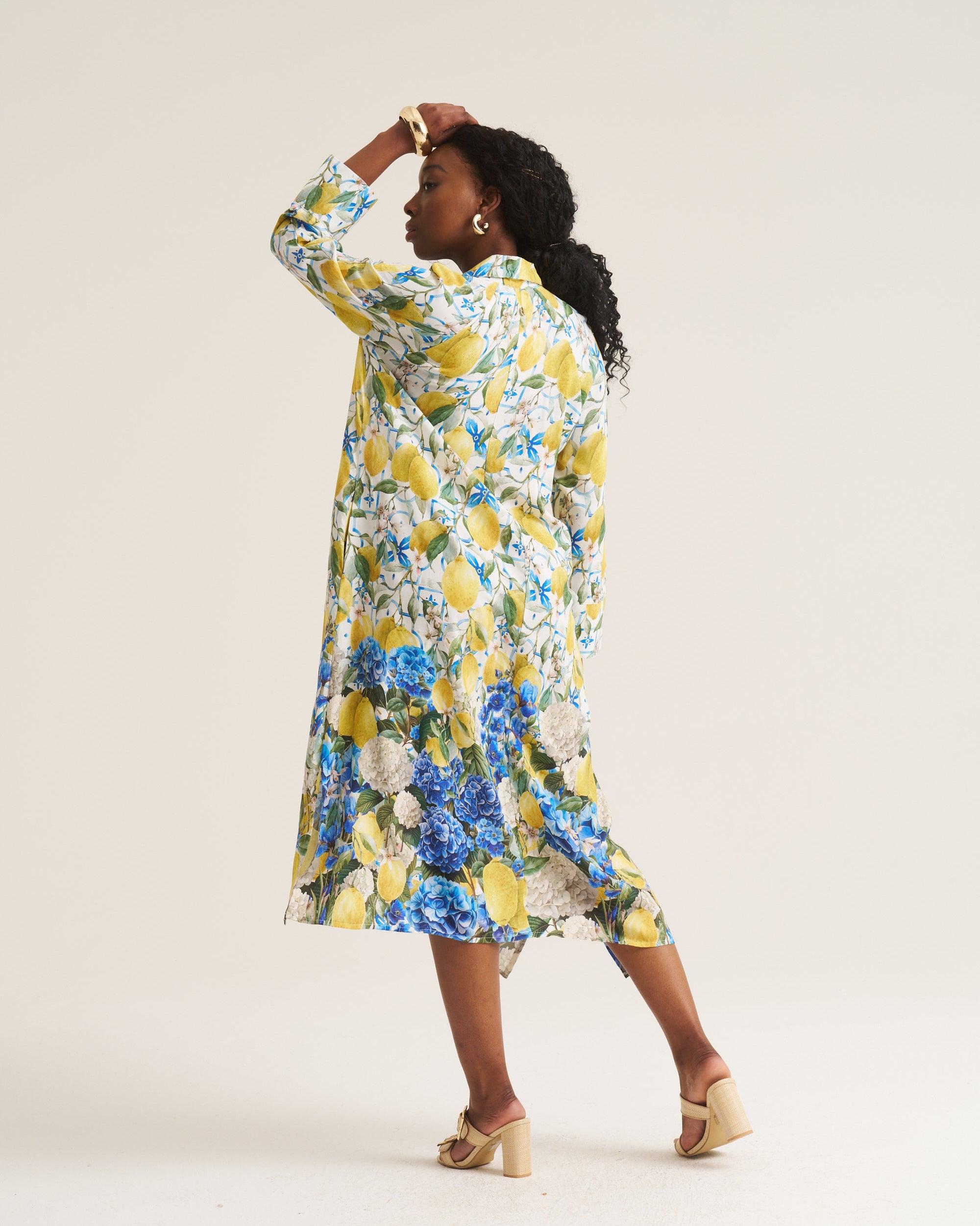 A woman modeling a long-sleeved midi dress with a vibrant yellow, blue, and white floral print, photographed from behind against a solid light beige studio background.