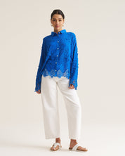 A woman modeling a vibrant blue long-sleeve button-up blouse with intricate floral lace embroidery and white wide-leg cropped pants, standing against a seamless off-white studio background.