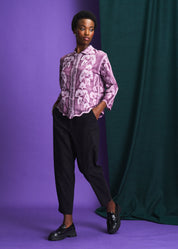 Person wearing a purple floral blouse and black pants against a purple and green backdrop