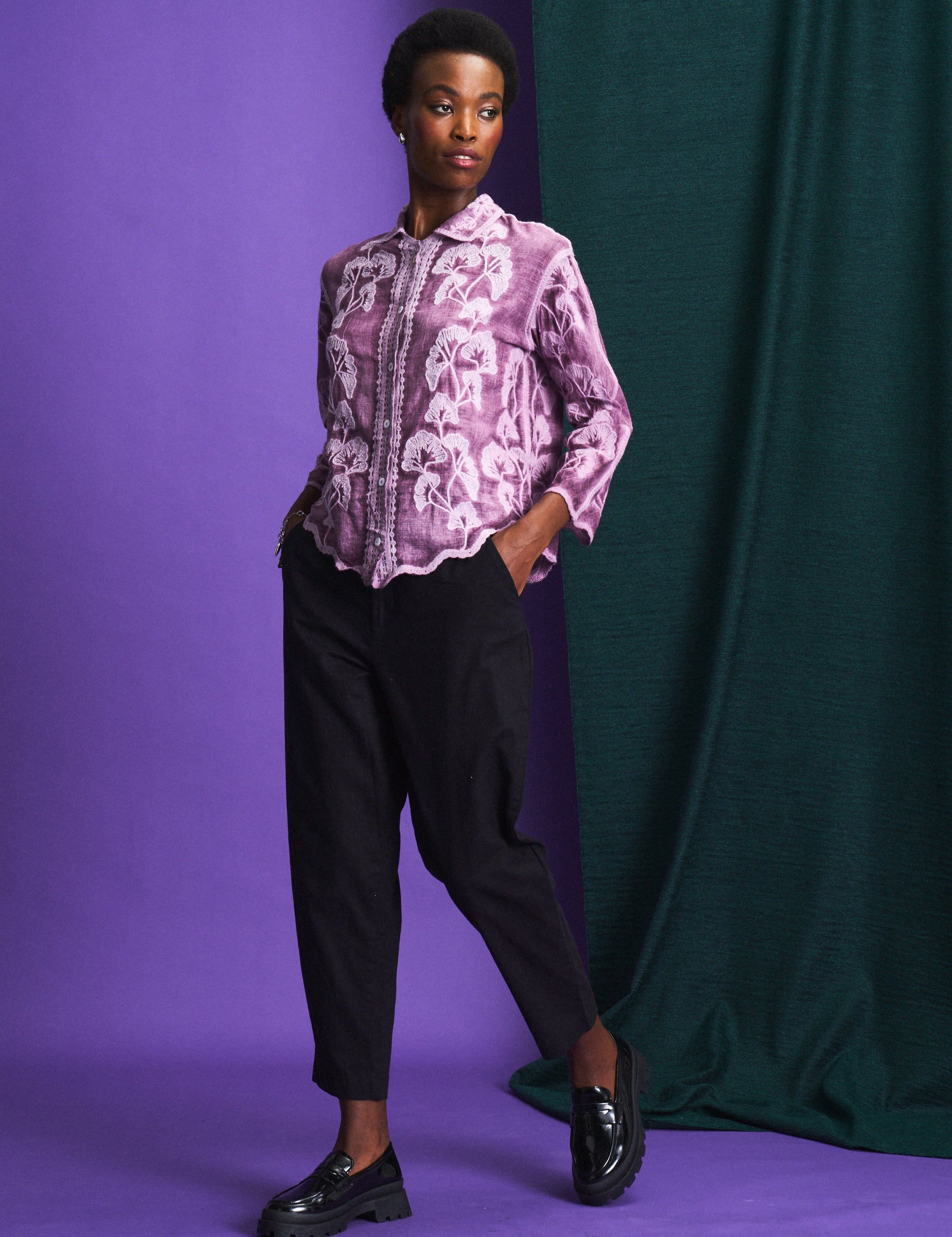 Person wearing a purple floral blouse and black pants against a purple and green backdrop