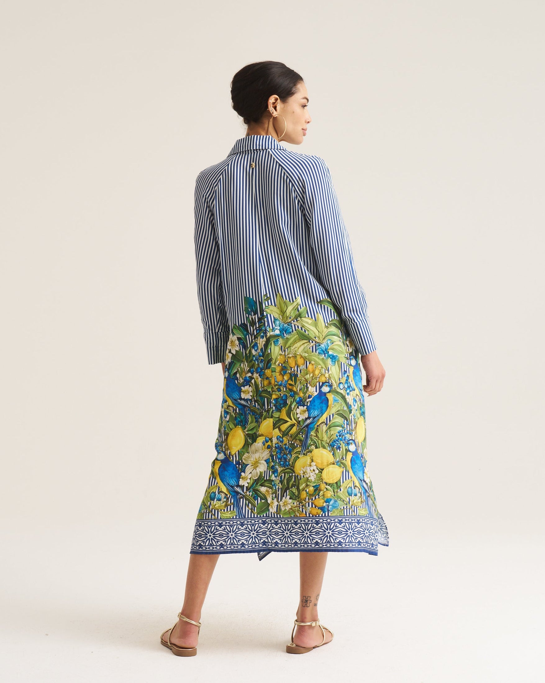 A woman is shown from behind wearing a blue and white striped long-sleeve shirt and a vibrant green, yellow, and blue floral print midi skirt with a blue patterned hem, set against a plain off-white studio background.