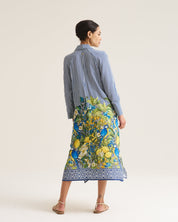 A woman is shown from behind wearing a blue and white striped long-sleeve shirt and a vibrant green, yellow, and blue floral print midi skirt with a blue patterned hem, set against a plain off-white studio background.