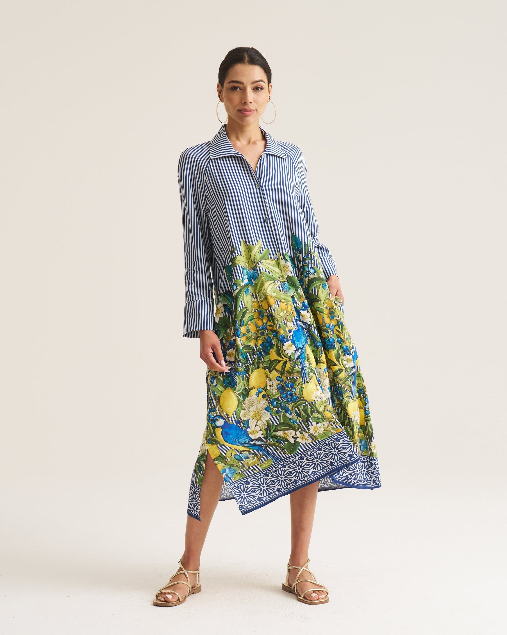 A woman modeling a long-sleeve midi dress with a blue and white striped upper bodice and a vibrant blue, green, and yellow floral print lower skirt, standing against a plain off-white studio background.