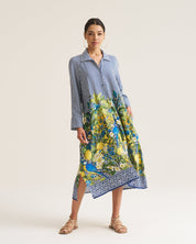 A woman modeling a long-sleeve midi dress with a blue and white striped upper bodice and a vibrant blue, green, and yellow floral print lower skirt, standing against a plain off-white studio background.