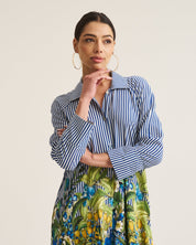 A woman modeling a long-sleeve shirt dress with blue and white vertical stripes on the top and a green, yellow, and blue tropical floral print on the skirt, set against a plain light beige background.