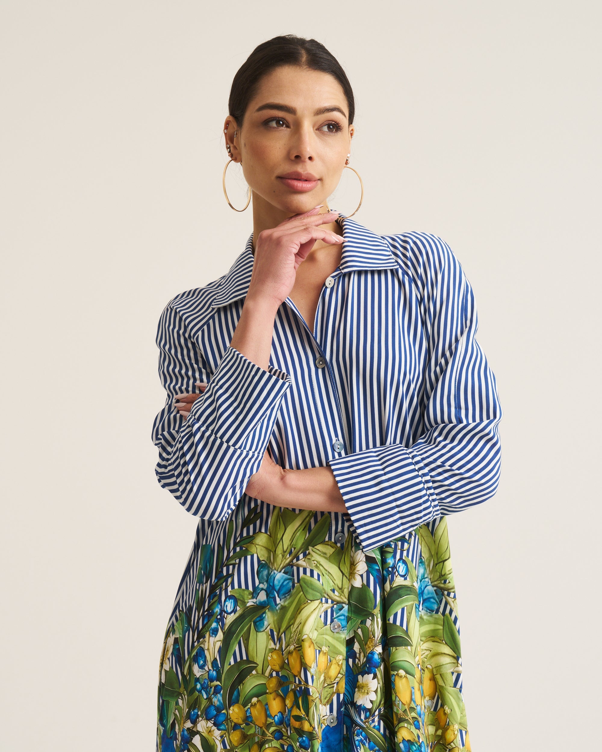 A woman modeling a long-sleeve shirt dress with blue and white vertical stripes on the top and a green, yellow, and blue tropical floral print on the skirt, set against a plain light beige background.