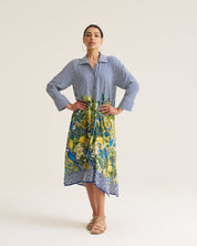 A woman modeling a long-sleeve shirt dress with a blue and white striped upper section and a colorful blue, yellow, and green floral print lower skirt, standing against a plain light beige studio background.