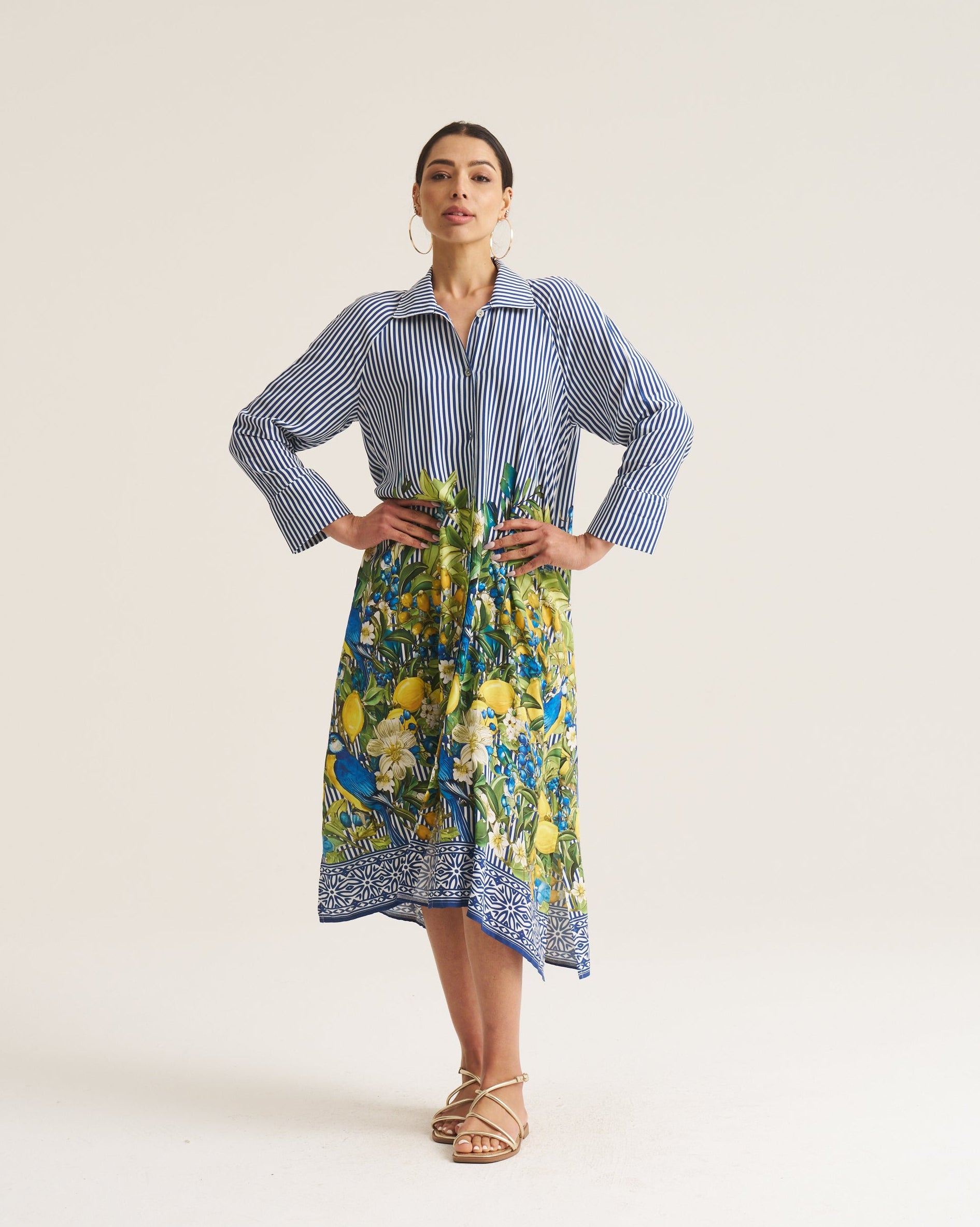 A woman modeling a long-sleeve shirt dress with a blue and white striped upper section and a colorful blue, yellow, and green floral print lower skirt, standing against a plain light beige studio background.