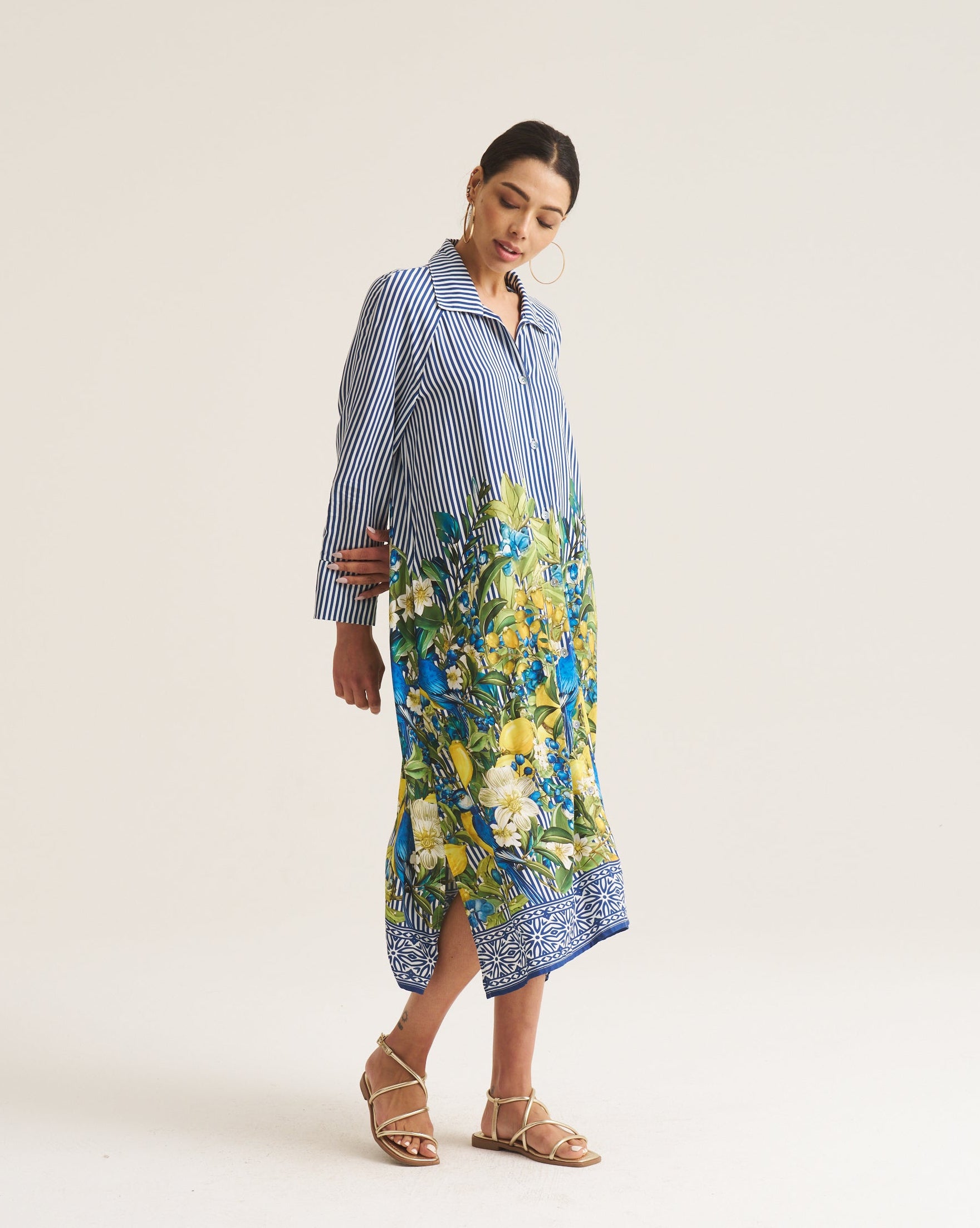 A woman modeling a long-sleeve midi shirt dress with a blue and white striped upper section and a vibrant blue, green, and yellow floral print lower section, paired with beige strappy sandals against a plain off-white studio background.