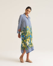 A woman modeling a long-sleeve midi shirt dress with a blue and white striped upper section and a vibrant blue, green, and yellow floral print lower section, paired with beige strappy sandals against a plain off-white studio background.