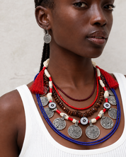 A close-up portrait of a woman wearing a layered bohemian-style necklace set with silver coin charms, red tassels, red and blue beaded strands, and brown beads, paired with a white ribbed tank top against a neutral beige wall.