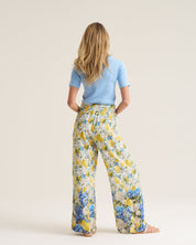 A woman stands with her back to the camera wearing wide-leg floral pants in yellow, blue, and white, paired with a light blue short-sleeve top, against a plain beige studio background.