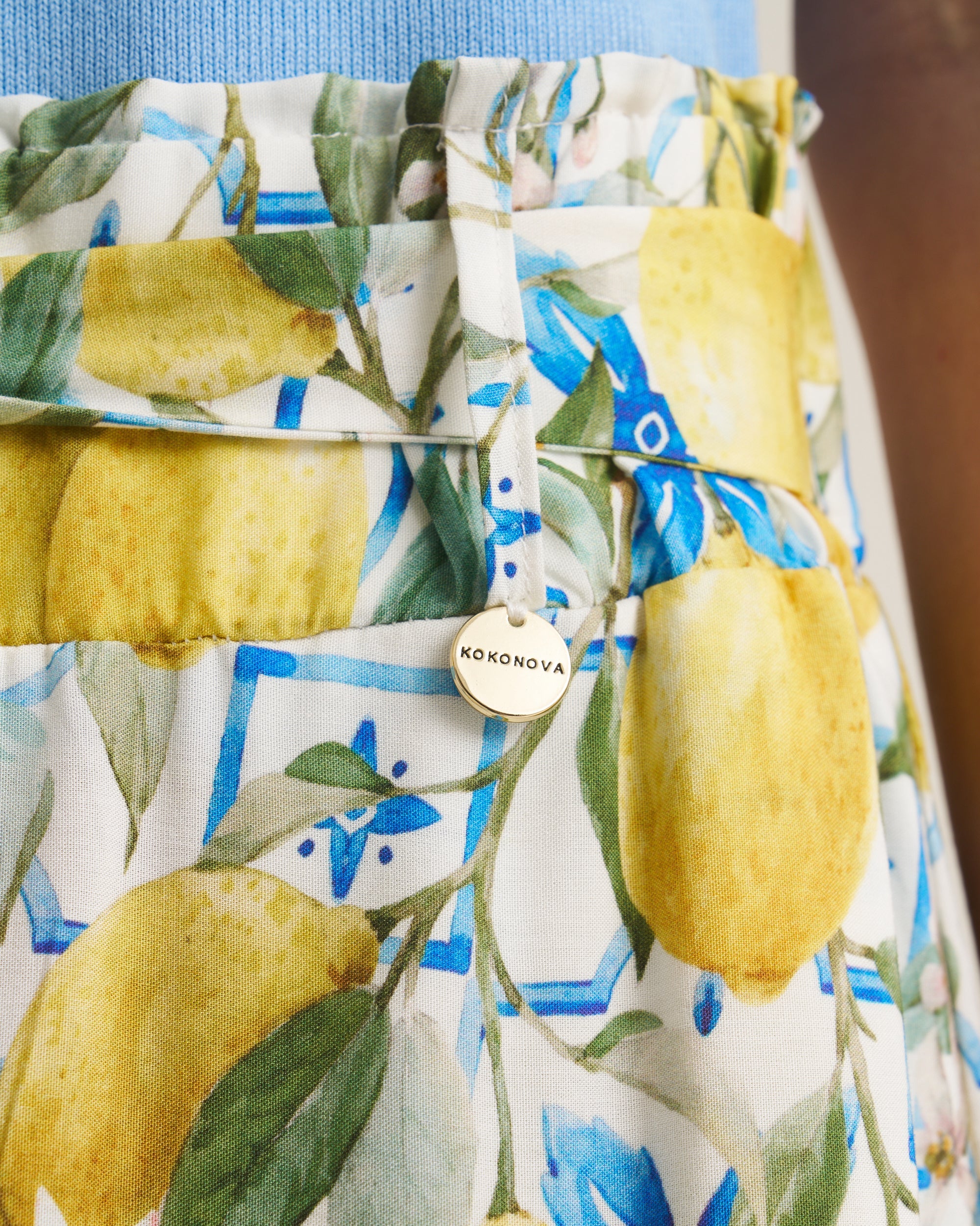 A close-up of a patterned fabric item, likely a skirt or apron, featuring a watercolor print of yellow lemons, green leaves, and blue geometric accents on a white background. A small round metal button with the brand name 'KOKONOVA' is visible.