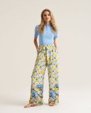 A woman modeling a light blue short-sleeve polo shirt and high-waisted, wide-leg trousers with a yellow, blue, and white floral print, standing against a solid light beige studio background.