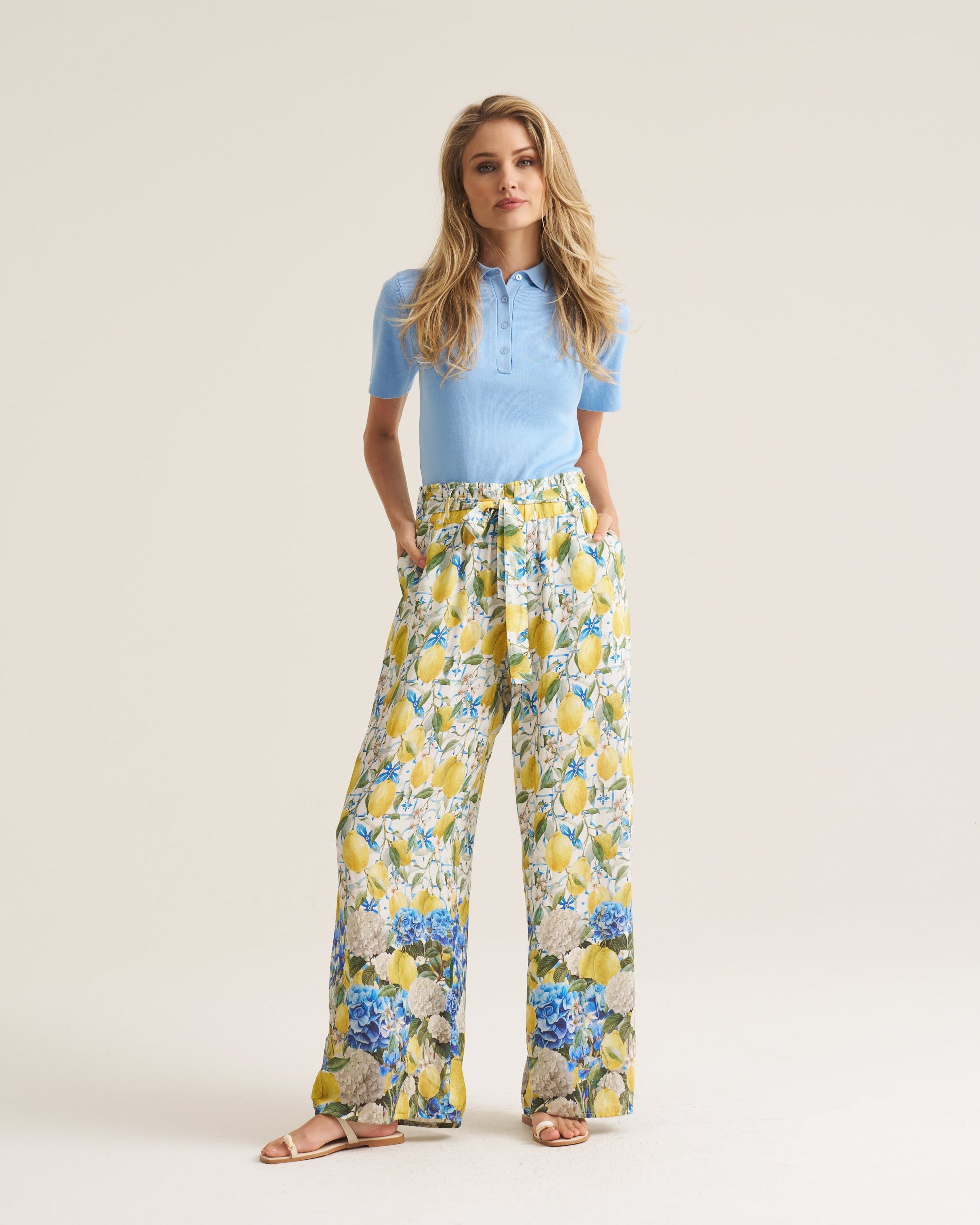 A woman modeling a light blue short-sleeve polo shirt and high-waisted, wide-leg trousers with a yellow, blue, and white floral print, standing against a solid light beige studio background.