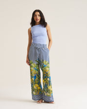 A woman modeling wide-leg trousers with a blue and white striped upper section and a vibrant green and yellow botanical print lower section, paired with a light blue sleeveless top, against a plain beige studio background.