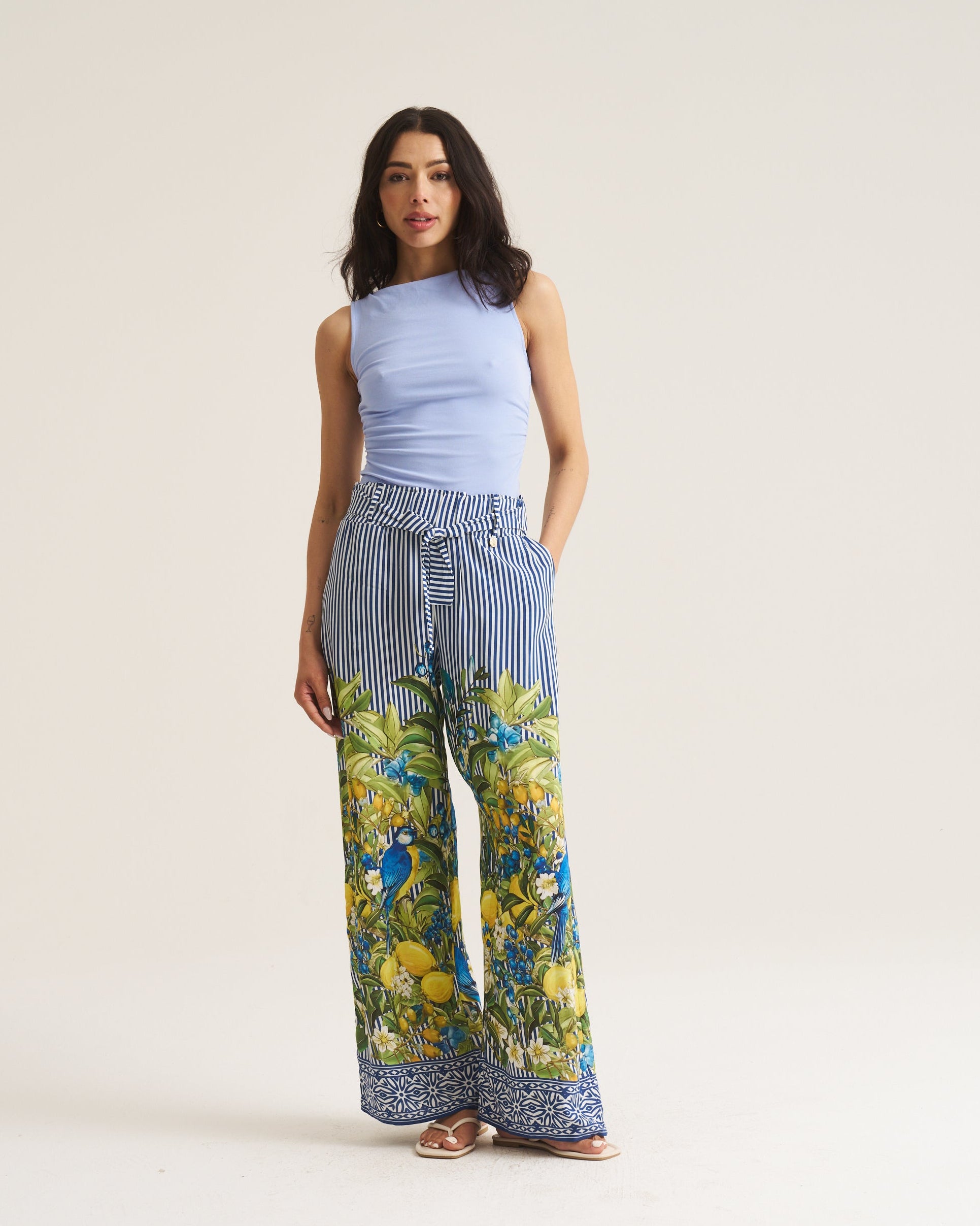 A woman modeling wide-leg trousers with a blue and white striped upper section and a vibrant green and yellow botanical print lower section, paired with a light blue sleeveless top, against a plain beige studio background.