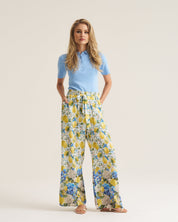 A woman stands against a plain light beige studio background wearing a light blue short-sleeve polo shirt and wide-leg trousers with a yellow, blue, and white floral print.