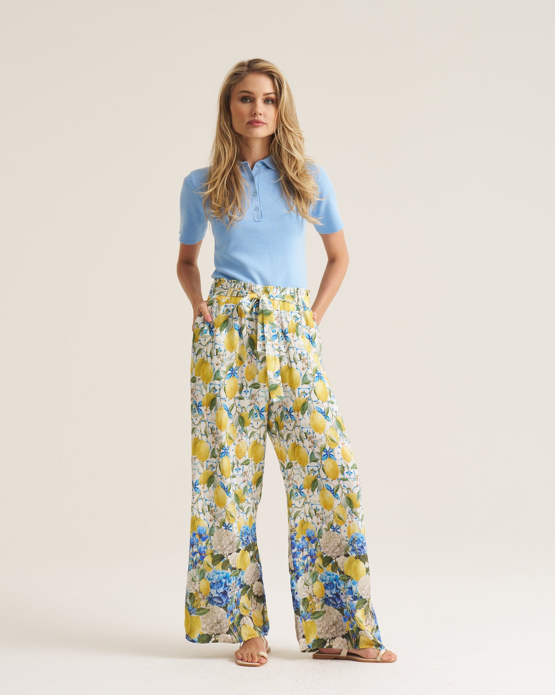 A woman stands against a plain light beige studio background wearing a light blue short-sleeve polo shirt and wide-leg trousers with a yellow, blue, and white floral print.