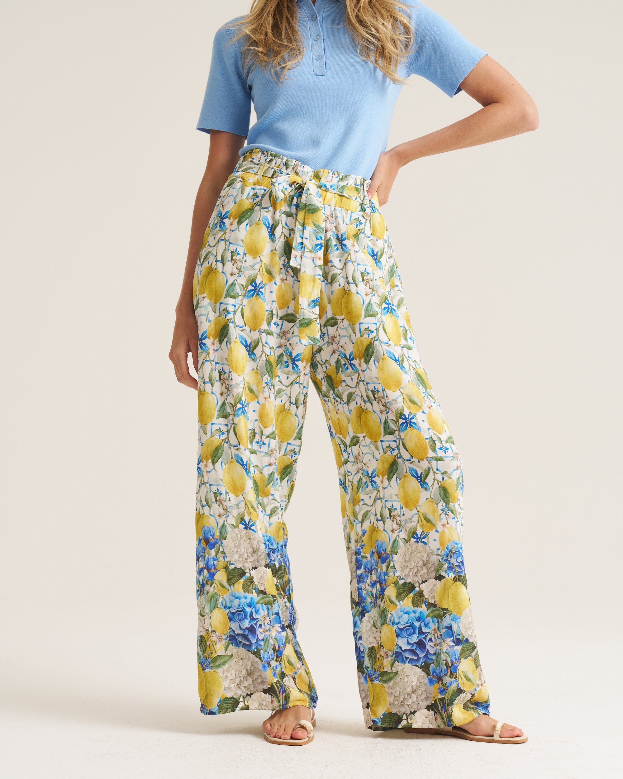 A model wearing high-waisted, wide-leg trousers with a yellow and blue floral print, paired with a light blue polo shirt and tan sandals, against a plain beige studio background.