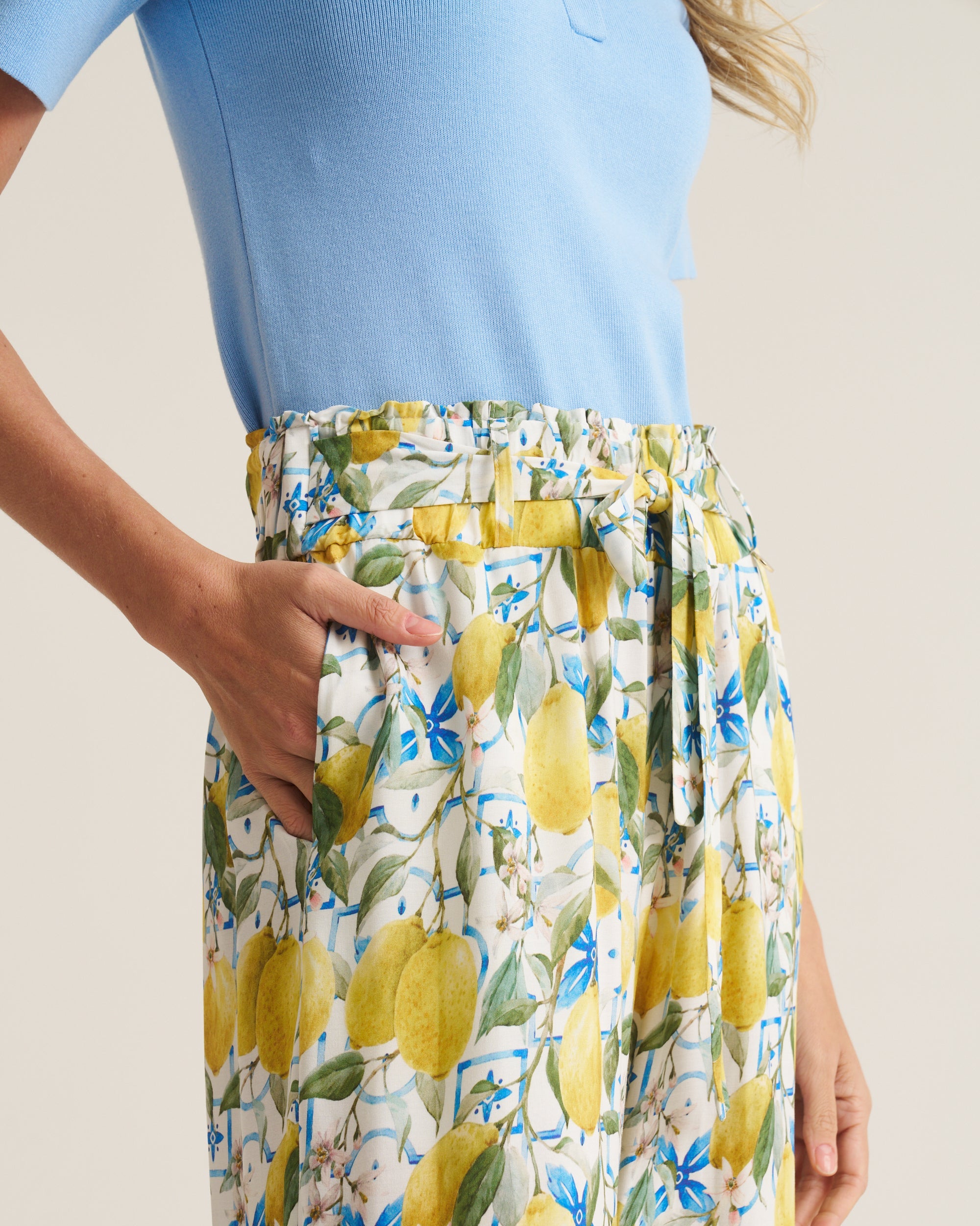 A close-up of a model wearing high-waisted, wide-leg trousers with a yellow lemon and blue floral print on a white background, paired with a light blue top.