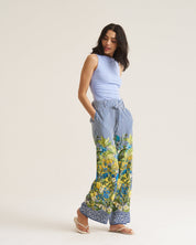 A woman modeling wide-leg pants with a blue and white striped upper section and a green and blue floral print lower section, paired with a light blue sleeveless top and beige sandals, against a plain off-white studio background.