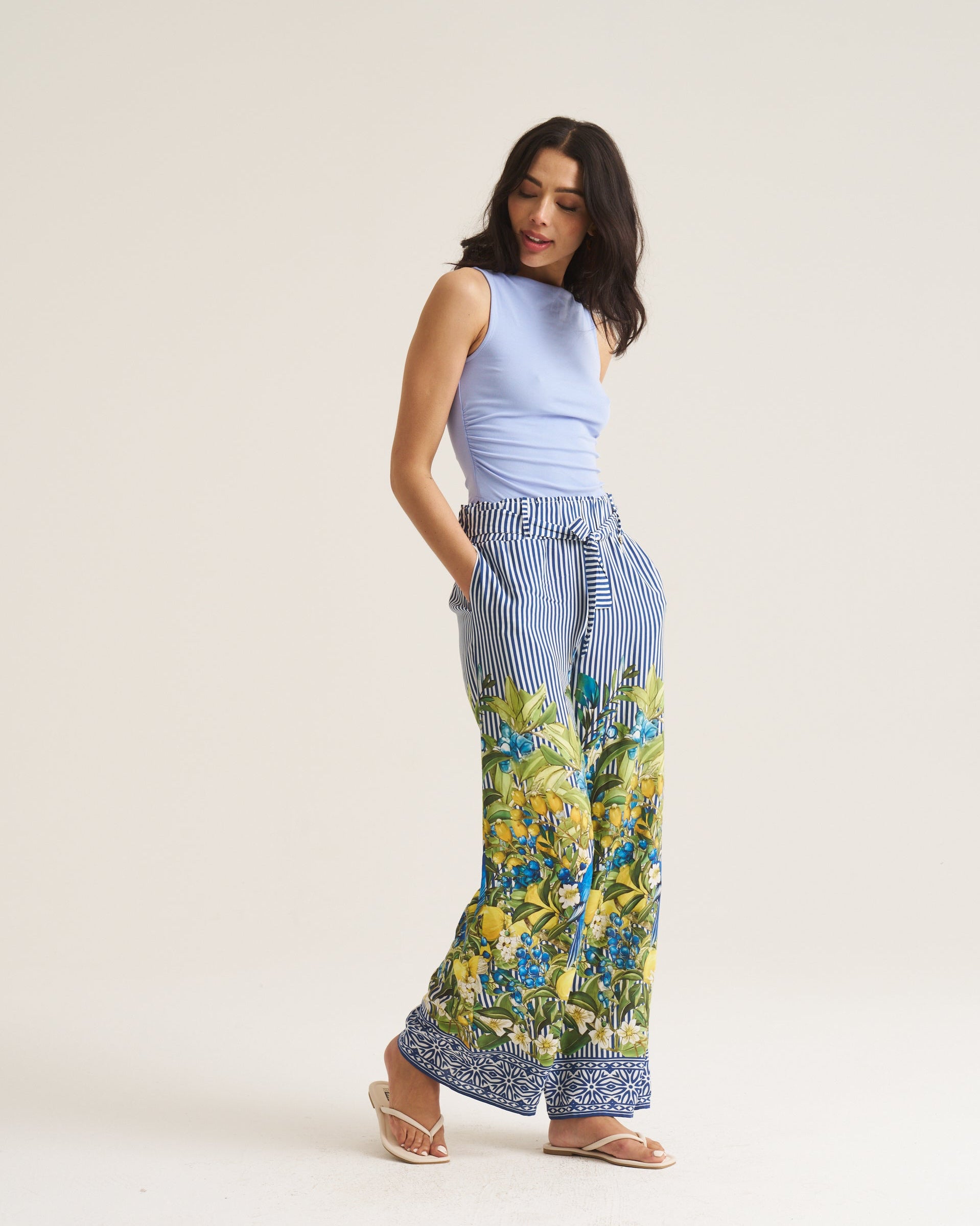 A woman modeling wide-leg pants with a blue and white striped upper section and a green and blue floral print lower section, paired with a light blue sleeveless top and beige sandals, against a plain off-white studio background.