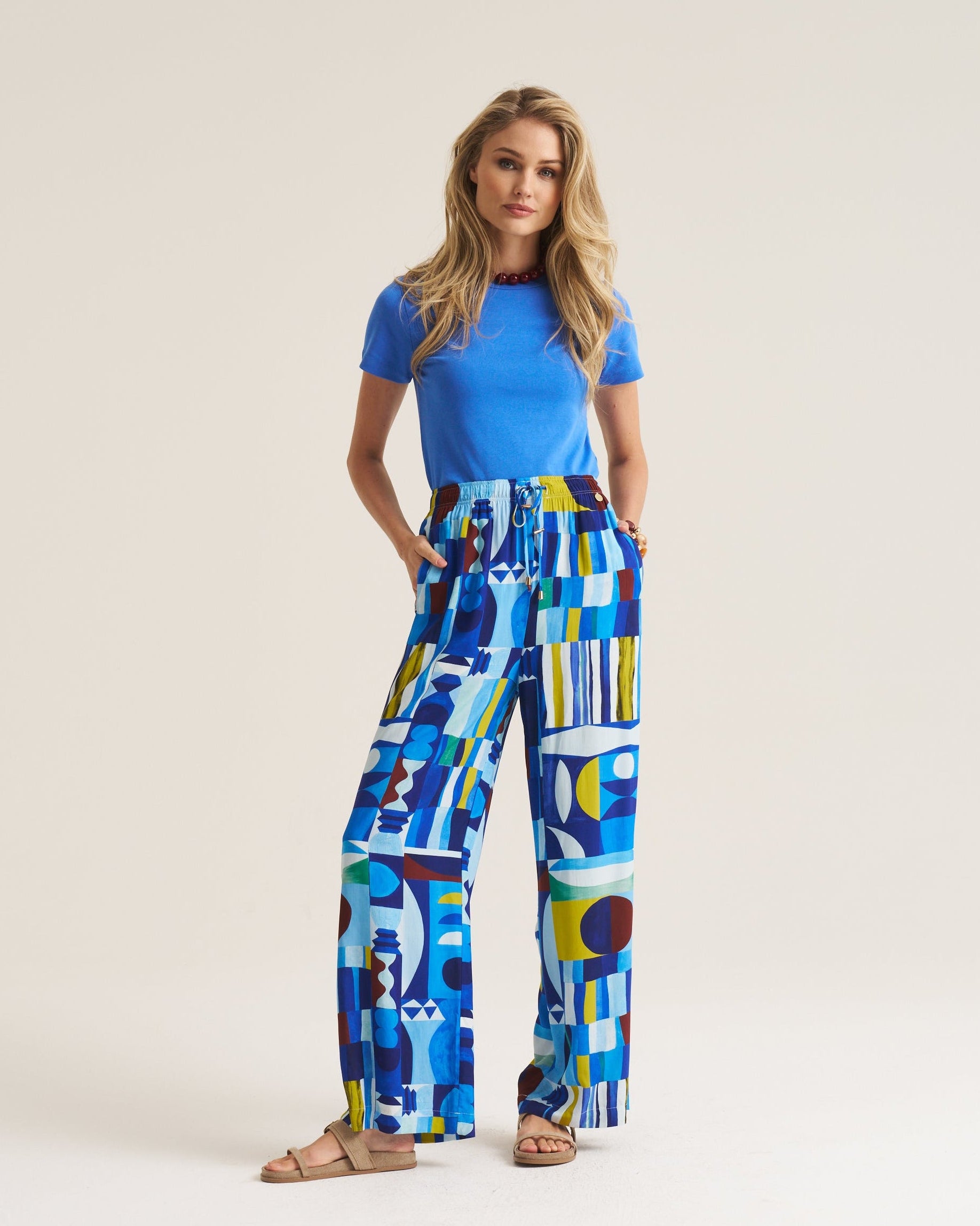 A woman modeling a bright blue short-sleeve top and high-waisted wide-leg trousers featuring a bold abstract geometric print in blue, yellow, white, and black, paired with tan flat sandals against a solid light beige background.