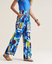 A model wearing high-waisted, wide-leg trousers with a colorful geometric abstract print in shades of blue, yellow, white, and brown, paired with a blue top and tan sandals.