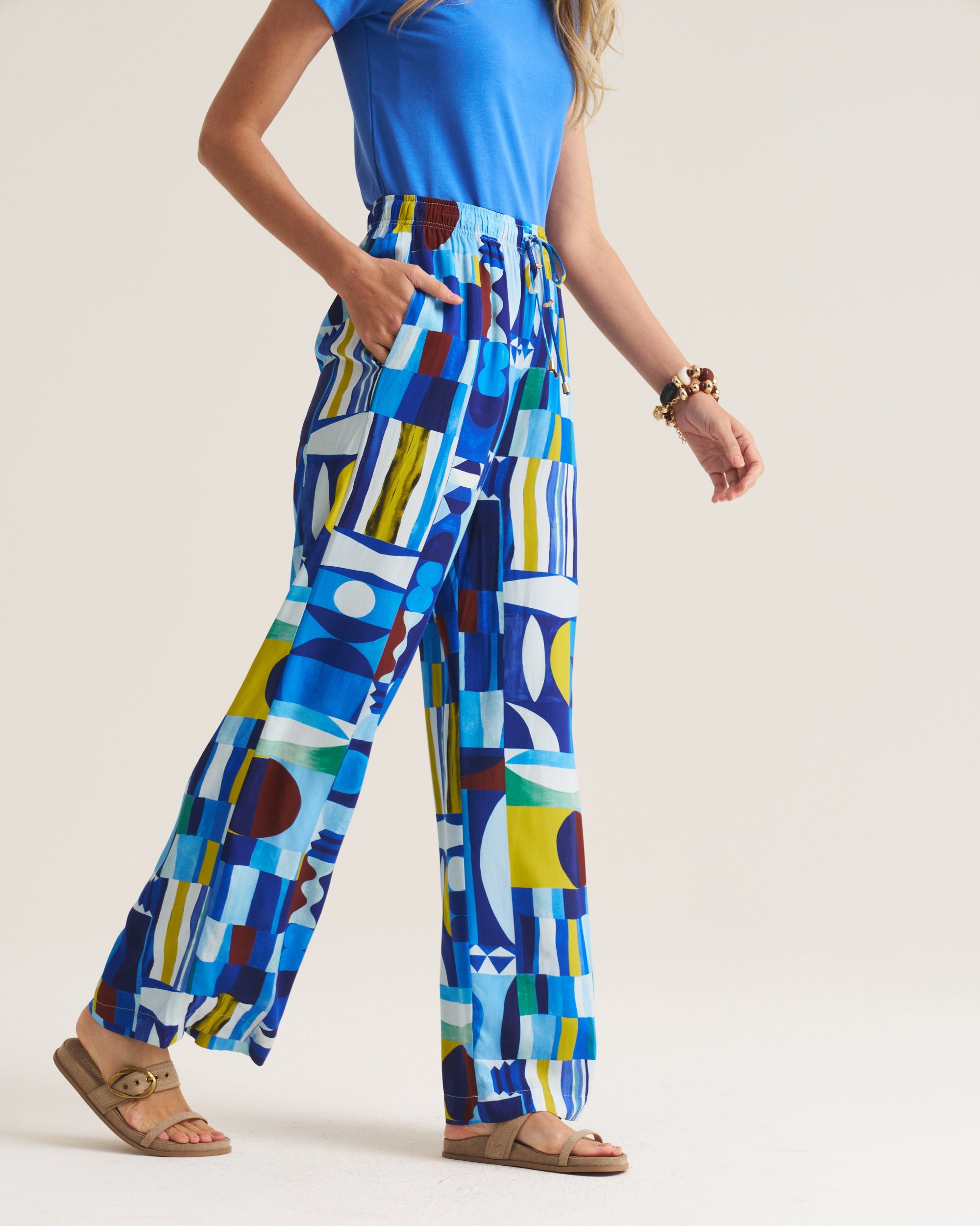A model wearing high-waisted, wide-leg trousers with a colorful geometric abstract print in shades of blue, yellow, white, and brown, paired with a blue top and tan sandals.