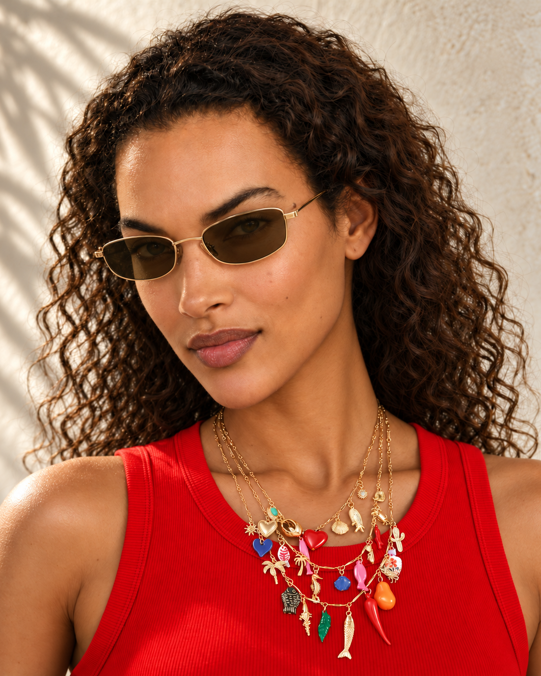 A woman with curly brown hair wearing gold-framed sunglasses with dark lenses and a red top, accessorized with layered gold necklaces featuring colorful charms, against a textured beige wall with soft leaf shadows.