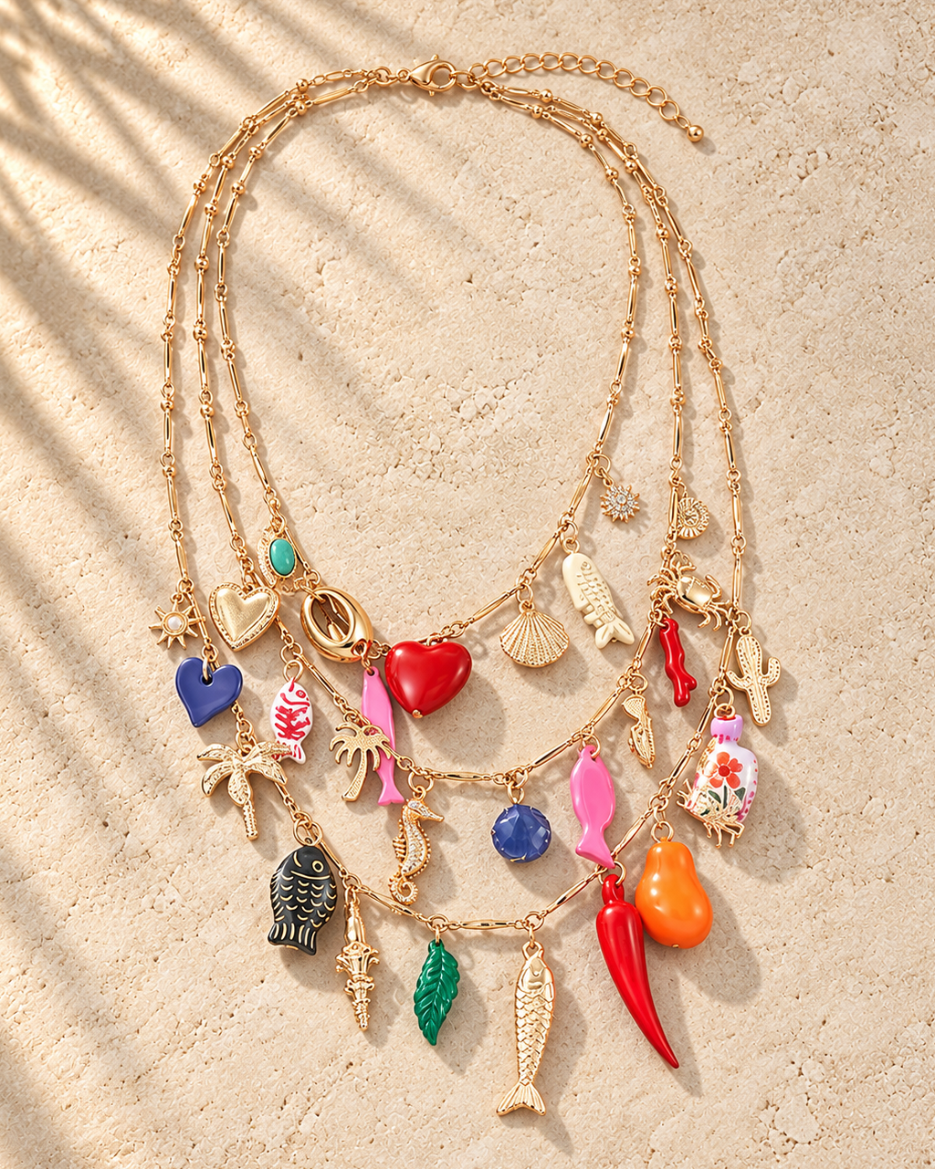 A gold multi-strand necklace with various colorful charms, including fish, hearts, seashells, and tropical motifs, displayed on a textured beige surface with soft palm leaf shadows.