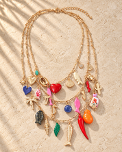 A gold multi-strand necklace with various colorful charms, including fish, hearts, seashells, and tropical motifs, displayed on a textured beige surface with soft palm leaf shadows.