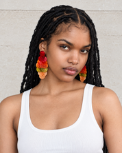 A woman with black dreadlocks wearing large, colorful flower-shaped earrings in red, orange, yellow, and green, paired with a white ribbed tank top against a light beige wall.