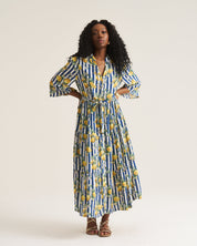 A woman modeling a long-sleeved maxi dress with a blue and white vertical stripe pattern and yellow lemon prints, paired with brown strappy sandals against a plain light beige studio background.