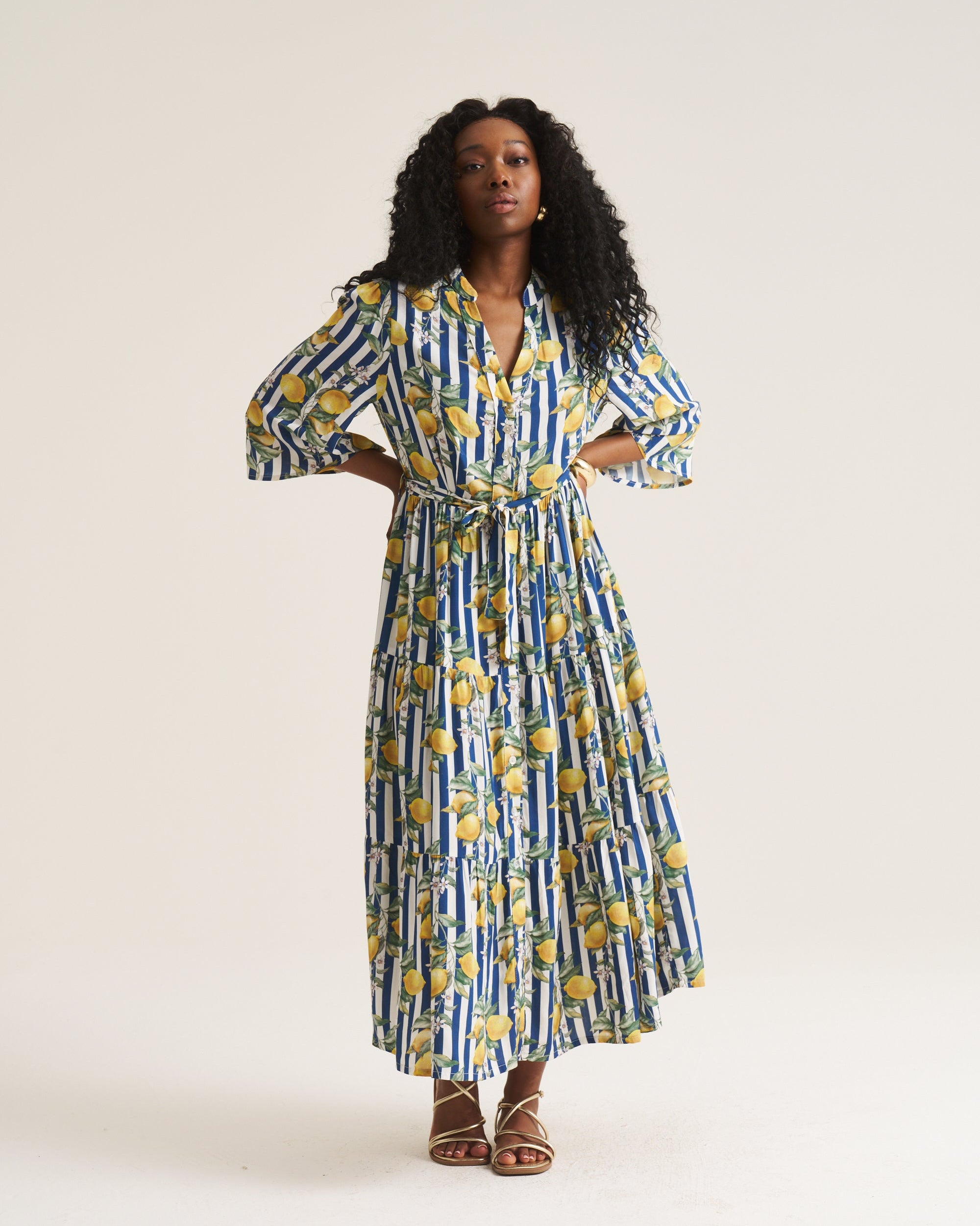 A woman modeling a long-sleeved maxi dress with a blue and white vertical stripe pattern and yellow lemon prints, paired with brown strappy sandals against a plain light beige studio background.