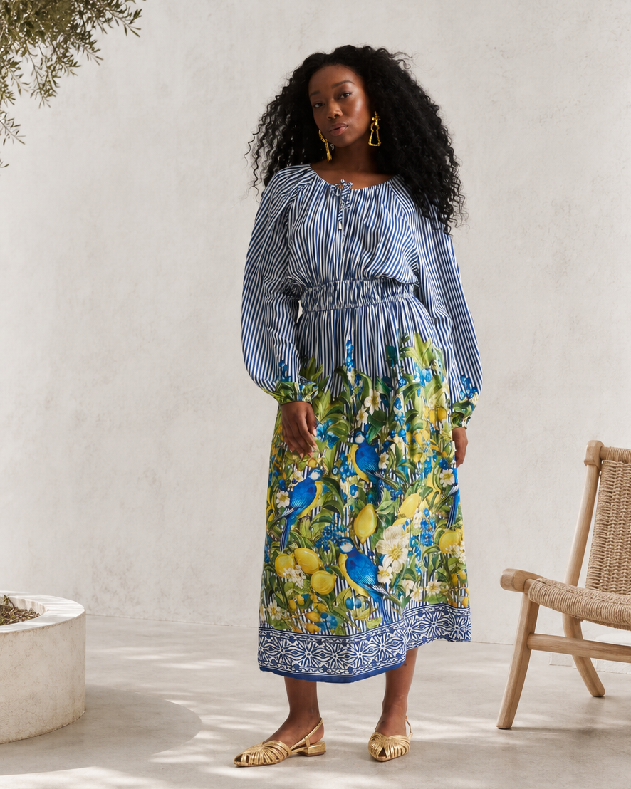 A woman stands in a bright, minimalist indoor patio setting wearing a long-sleeve midi dress with a blue and white striped bodice and a vibrant blue, green, and yellow floral print skirt. She is wearing gold flat sandals and gold hoop earrings.
