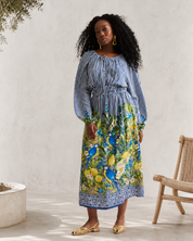 A woman stands in a bright, minimalist indoor patio setting wearing a long-sleeve midi dress with a blue and white striped bodice and a vibrant blue, green, and yellow floral print skirt. She is wearing gold flat sandals and gold hoop earrings.