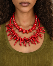A woman with curly dark hair wearing a bright red statement necklace and an olive green ribbed tank top, photographed against a light textured wall.